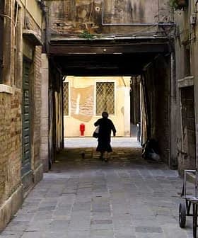 Un individu marchant dans une ruelle étroite avec des murs de briques et un chat qui le suit. - Olive Oil Times