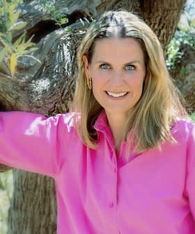 Donna con i capelli lunghi che indossa una maglietta rosa, appoggiata a un ulivo con un sorriso. - Olive Oil Times