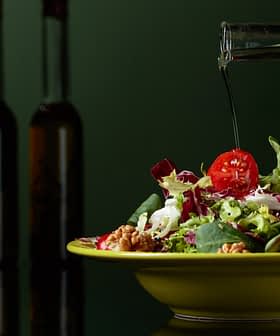 Aceite de oliva vertido sobre una ensalada fresca en un recipiente amarillo con varias verduras. Olive Oil Times