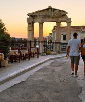 Una pareja caminando por una calle cerca de ruinas antiguas al atardecer, con mesas de comedor al aire libre visibles. Olive Oil Times