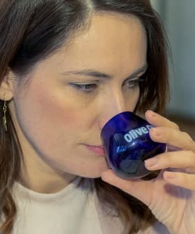 Donna con i capelli castani che tiene vicino al viso un bicchiere da degustazione blu con la scritta "Olio d'oliva". Olive Oil Times