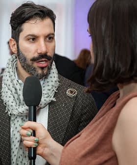 Man met een baard, een jas met patroon en een sjaal, die tijdens een interview in een microfoon spreekt. Olive Oil Times