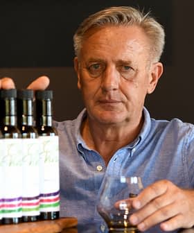 Gri saçlı adam, masanın üzerinde sıralanmış zeytinyağı şişelerinin önünde bir bardak tutuyor. - Olive Oil Times
