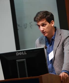 Ein Mann in einem grauen Blazer spricht während einer Präsentation an einem Podium mit einem Dell-Monitor. - Olive Oil Times
