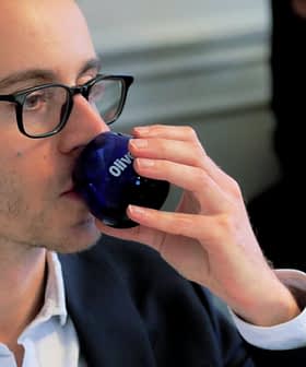眼鏡をかけた男性が、「オリーブ」と書かれた青いグラスから飲んでいます。 - Olive Oil Times