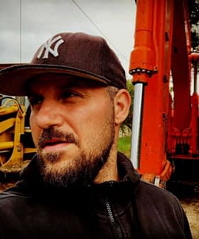 Čovjek s kapom stoji blizu narančastog bagera na gradilištu. - Olive Oil Times