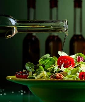 Uma mão despejando azeite de oliva de uma garrafa de vidro sobre uma salada em uma tigela verde com tomates cereja e verduras. - Olive Oil Times