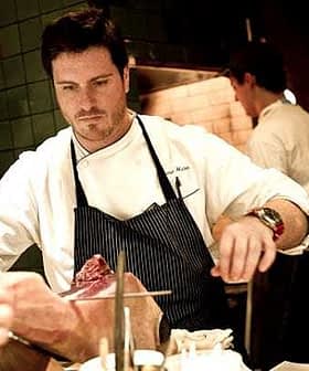 Chef en blouse blanche et tablier préparant un morceau de viande dans un décor de cuisine. - Olive Oil Times