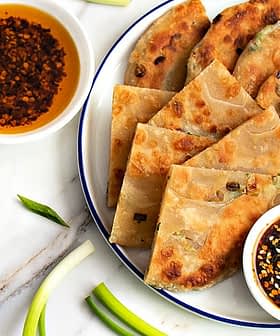 Ein Teller mit Frühlingszwiebelpfannkuchen, angerichtet mit zwei Schüsseln Dip-Saucen und Frühlingszwiebeln auf einer Marmoroberfläche. - Olive Oil Times
