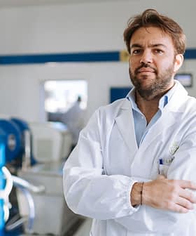 Man wearing a laboratory coat standing with arms crossed in a facility with machinery in the background. - Olive Oil Times