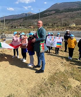 Gruppo di bambini che indossano berretti blu durante una cerimonia di piantagione di ulivi con una bandiera italiana. Olive Oil Times