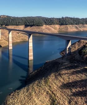 Un puente que se extiende sobre un cuerpo de agua con pilares de soporte y un paisaje circundante. Olive Oil Times