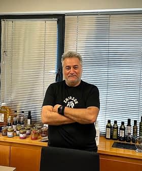 Un homme aux cheveux gris portant un t-shirt noir debout dans un bureau avec des produits à base d'huile d'olive exposés sur une étagère. - Olive Oil Times