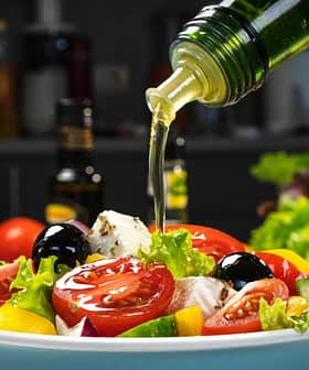 Olijfolie wordt over een verse salade met tomaten, sla en olijven in een kom gegoten. Olive Oil Times