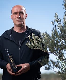 Mann steht im Freien und hält eine Flasche Olivenöl neben einem Olivenbaum. - Olive Oil Times