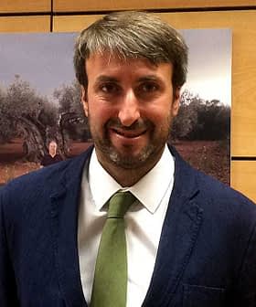 Homem vestindo um terno escuro e uma gravata verde, em pé em frente a um fundo com oliveiras. - Olive Oil Times