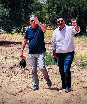 Twee mannen lopen door een olijfgaard, de een zwaait en de ander beschermt zijn ogen tegen de zon. Olive Oil Times