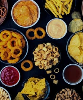Una variedad de bocadillos que incluyen papas fritas, pretzels y palomitas de maíz dispuestos en tazones sobre una superficie oscura. Olive Oil Times