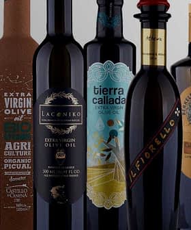 Una collezione di bottiglie di olio d'oliva con etichette e disegni distinti, disposte in fila. - Olive Oil Times