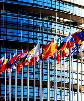 Várias bandeiras de países europeus exibidas em frente a um moderno edifício de vidro. - Olive Oil Times