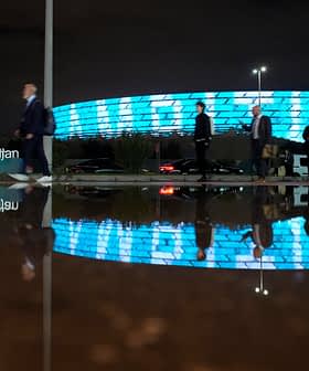 Ομάδα ανθρώπων που περπατά δίπλα από ένα σύγχρονο κτήριο με φωτισμένη μπλε πρόσοψη τη νύχτα, που αντανακλάται σε μια λακκούβα. - Olive Oil Times