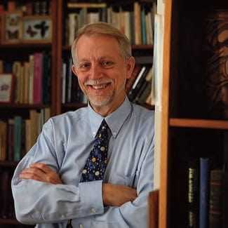A man with gray hair and glasses smiling while standing in front of bookshelves filled with books. - Olive Oil Times