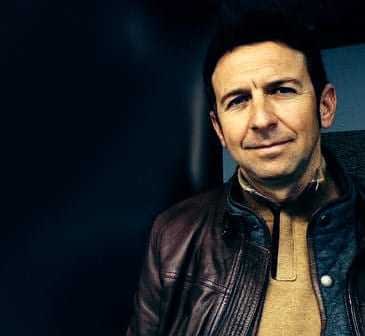 A man wearing a brown leather jacket with a neutral expression against a dark background. - Olive Oil Times