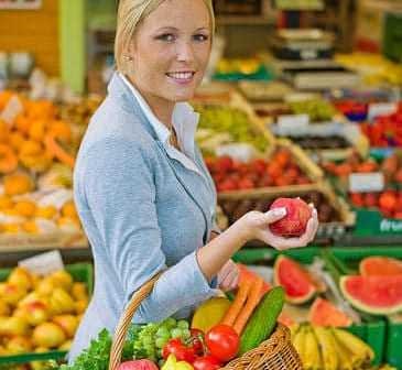 Woman in a gray blazer holding a red apple while carrying a basket of fresh fruits and vegetables. - Olive Oil Times