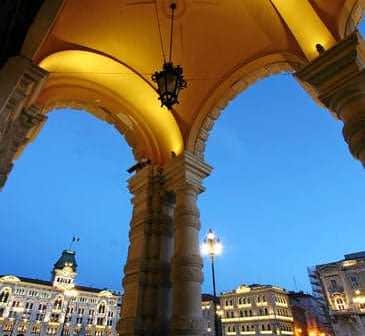 Archway with decorative columns and lanterns, overlooking a city square in Trieste during twilight. - Olive Oil Times