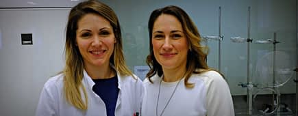 Two women smiling in a laboratory environment, one wearing a lab coat and the other in a white sweater. - Olive Oil Times
