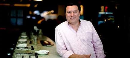 A man wearing a checkered shirt stands at a bar with a neutral expression. - Olive Oil Times