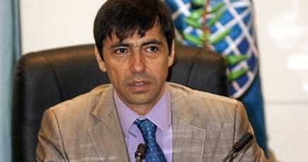 A man wearing a light-colored suit and blue tie sitting at a conference table with a serious expression. - Olive Oil Times