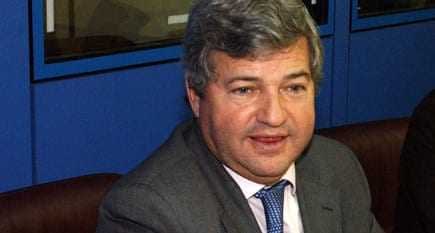 会議またはミーティング中にテーブルに座っている、グレーのスーツと青いネクタイを着用した男性。 - Olive Oil Times