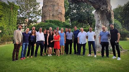 A group of individuals posing together for a photo in an outdoor area with greenery and a stone structure. - Olive Oil Times