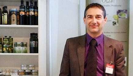 A man in a suit standing in front of a display of olive oil products on shelves. - Olive Oil Times