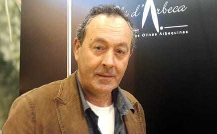 Man with dark hair and a light shirt, wearing a brown jacket, standing in front of a dark background. - Olive Oil Times