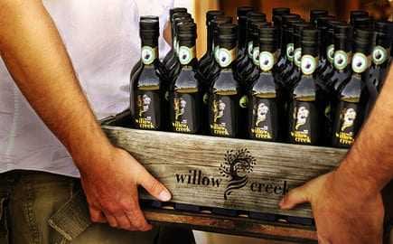 A wooden crate filled with bottles of Willow Creek olive oil held by two people. - Olive Oil Times