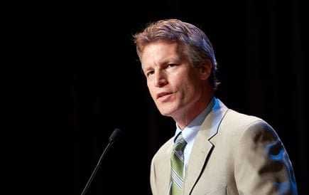 A man in a light suit and green tie speaking at a podium with a microphone. - Olive Oil Times