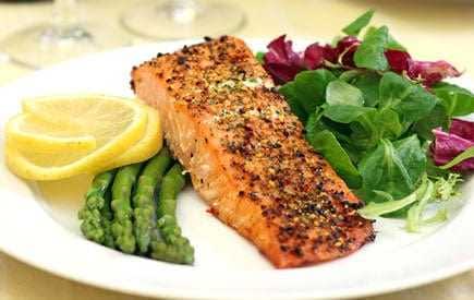 Grilled salmon fillet served with asparagus, lemon slices, and mixed salad greens on a white plate. - Olive Oil Times
