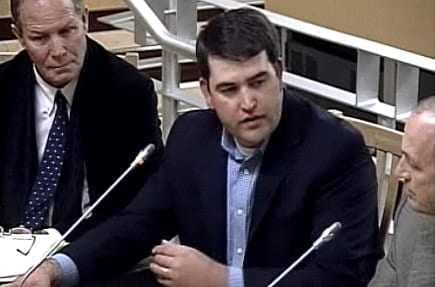 A man in a dark suit speaking during a meeting with two other men seated nearby. - Olive Oil Times