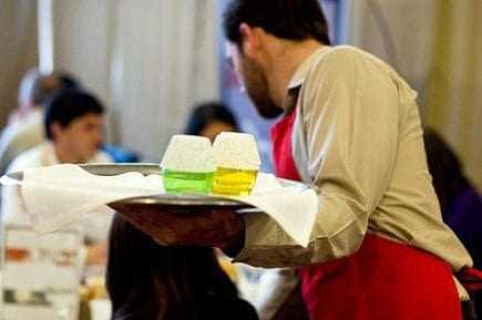 Waiter in a red apron holding a tray with two glasses of colored drinks at a dining event. - Olive Oil Times