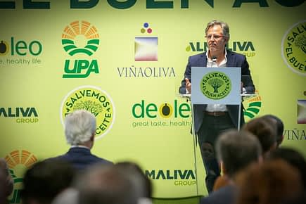 A speaker presenting at an olive oil industry event with a green backdrop featuring various logos. - Olive Oil Times