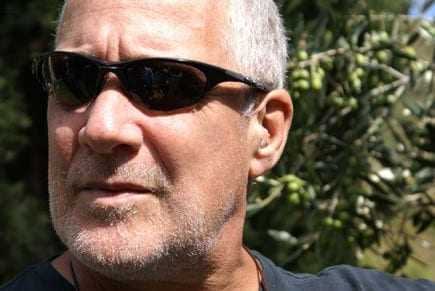 Man with short gray hair wearing sunglasses, with olive trees in the background. - Olive Oil Times