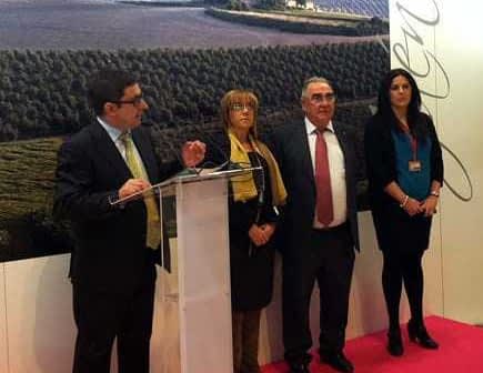 Four individuals standing at a podium during a presentation at an event, with a backdrop of olive fields. - Olive Oil Times