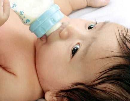 Infant with dark hair drinking from a blue baby bottle while lying down. - Olive Oil Times