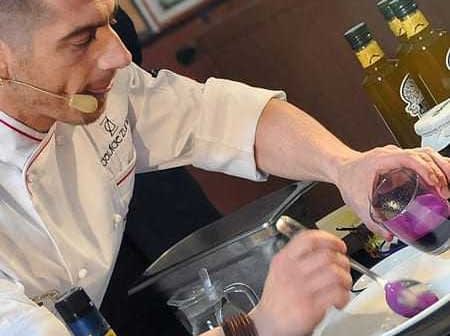 Chef in a white coat preparing a dish with olive oil and colorful ingredients on a countertop. - Olive Oil Times