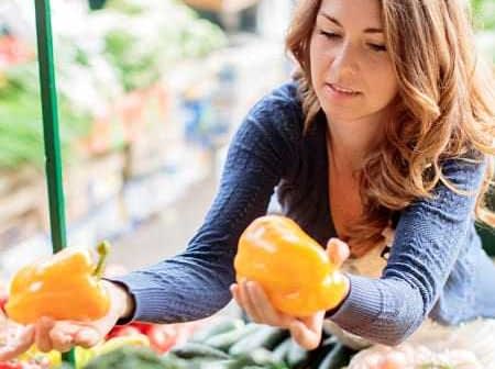 Woman examining yellow bell peppers while shopping at a market stall filled with vegetables. - Olive Oil Times