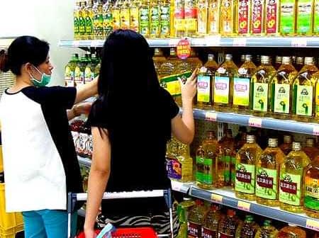 Two individuals shopping for cooking oils on a supermarket shelf filled with various oil bottles. - Olive Oil Times