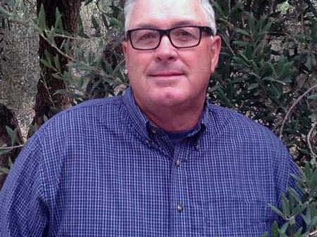 Man wearing glasses and a blue checkered shirt standing among olive trees. - Olive Oil Times