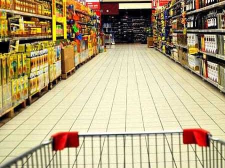 View of a supermarket aisle with shelves stocked with various products and a shopping cart in the foreground. - Olive Oil Times